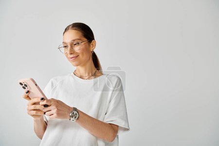 Eine junge Frau mit Brille hält ein Handy vor grauem Hintergrund.