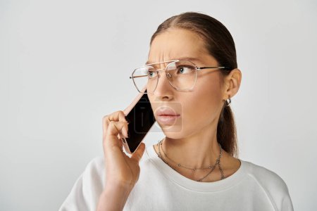 Una joven con una camiseta blanca y gafas hablando en un teléfono celular sobre un fondo gris.
