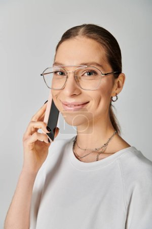 A young woman in a white t-shirt and glasses engaging in a conversation on a cell phone against a grey background.