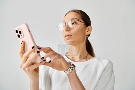 Eine stylische junge Frau mit Brille hält ein Handy vor grauem Hintergrund.