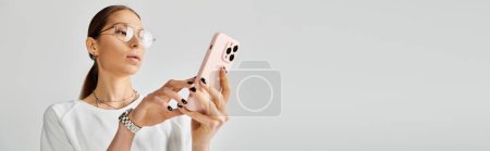Photo for A young woman in a white t-shirt and glasses holding a pink smartphone on a grey background. - Royalty Free Image