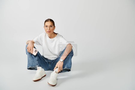 Photo for A young woman in a white t-shirt and glasses is sitting on the ground while holding a smartphone - Royalty Free Image