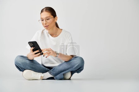 A young woman in a white t-shirt and glasses sits on the floor, engrossed in her cell phone.