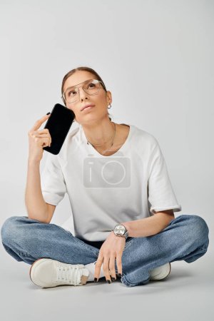Téléchargez les photos : Une jeune femme portant un t-shirt blanc et des lunettes est assise sur le sol, absorbée par son téléphone portable.. - en image libre de droit