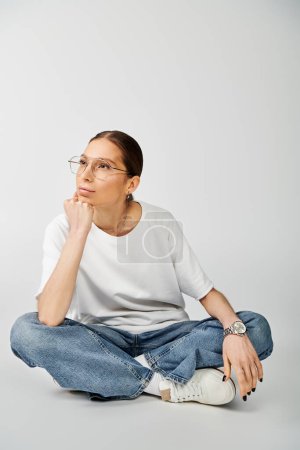 Eine junge Frau in weißem T-Shirt und Brille sitzt auf dem Boden, das Kinn in der Hand, verloren in Gedanken.