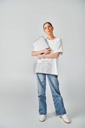 Téléchargez les photos : Une jeune femme se tient debout avec confiance avec un ordinateur portable, portant un t-shirt blanc et des lunettes sur un fond gris. - en image libre de droit