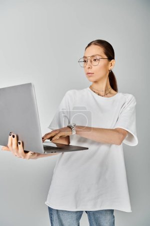 Una joven mujer sostiene con confianza un portátil en la mano, con una camiseta blanca y gafas sobre un fondo gris.