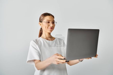 A young woman in a white t-shirt and glasses holding a laptop computer in her hands on a grey background.
