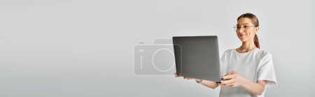 Téléchargez les photos : Une jeune femme en t-shirt blanc et lunettes tenant un ordinateur portable dans ses mains sur un fond gris. - en image libre de droit