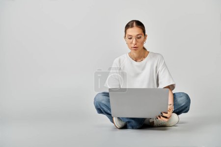 Téléchargez les photos : Une jeune femme dans un t-shirt blanc et des lunettes assises sur le sol, absorbées dans son ordinateur portable sur un fond gris. - en image libre de droit