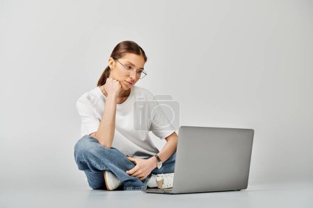 A young woman in a white t-shirt and glasses sits on the floor, focused on her laptop screen against a grey background.
