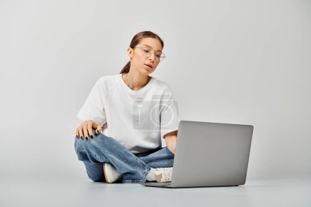 A young woman in a white t-shirt and glasses sits on the floor, focused on her laptop screen, engaged in work or study.