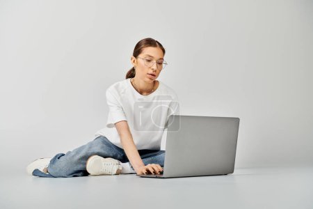 Eine junge Frau in weißem T-Shirt und Brille sitzt auf dem Fußboden und bedient ihren Laptop vor grauem Hintergrund..