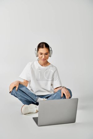 Foto de Mujer joven en camiseta blanca y gafas se sienta en el suelo con el ordenador portátil, centrado en los auriculares. - Imagen libre de derechos