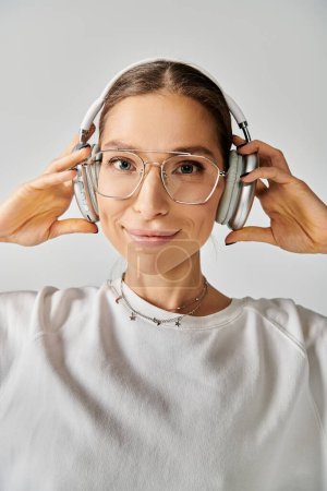 Photo for A young woman wearing glasses and headphones, listening intently on a grey background. - Royalty Free Image