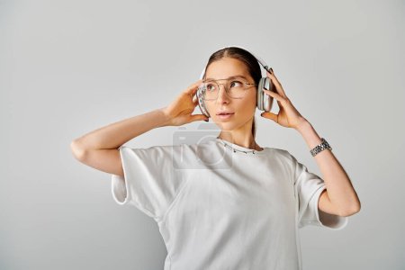 A woman in glasses holds a headphones to her ears, exuding intelligence and sophistication in a white t-shirt on a grey background.