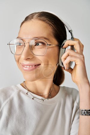 A young woman in a white t-shirt and glasses holding a headset on a grey background.