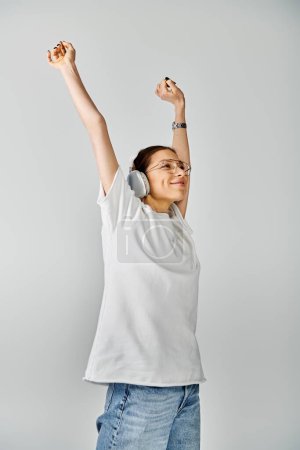 A young woman in a white shirt and glasses jubilantly lifts her arms against a grey background.