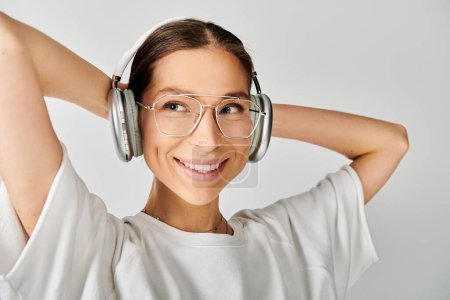 A young woman with glasses and headphones smiles contently against a grey background.
