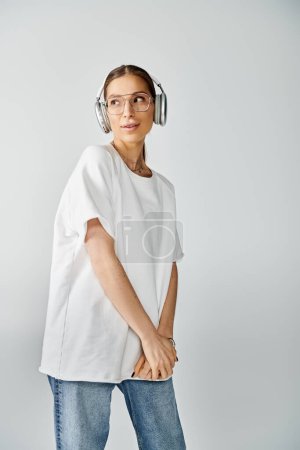 Une jeune femme en t-shirt blanc écoute de la musique à travers des écouteurs sur un fond gris, respirant le calme.
