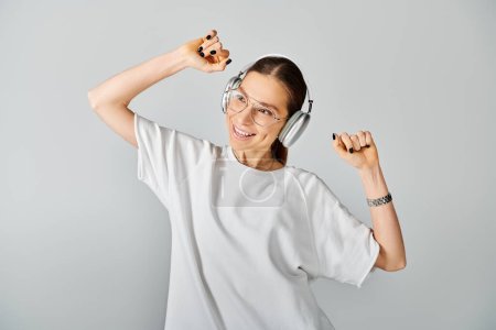 Eine junge Frau in weißem T-Shirt und Brille hält einen Kopfhörer vor grauem Hintergrund..