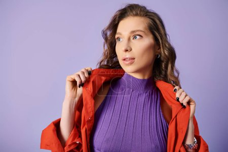 A stylish young woman in a purple top holding a vibrant jacket against a purple background.