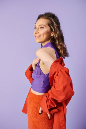 A stylish young woman standing out in a vibrant orange skirt and purple top against a purple background.