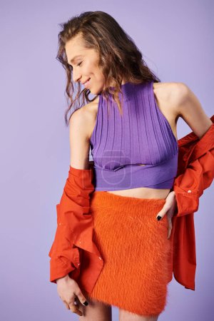 Une jeune femme élégante se distingue dans un haut violet et une jupe orange sur un fond violet vibrant.