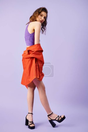 A stylish young woman stands out in a vibrant orange skirt and purple top against a matching background.