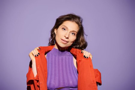 A stylish young woman stands out in a purple shirt and vibrant orange jacket against a purple background.