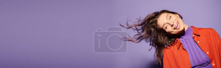 A stylish young woman in vibrant orange attire stands against a purple background, her hair gracefully blowing in the wind.