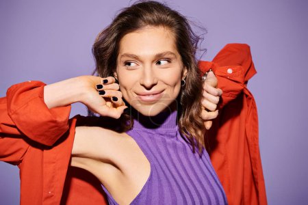 A stylish young woman wearing a purple top carefully holds a vibrant red jacket, creating a striking color contrast on a purple background.