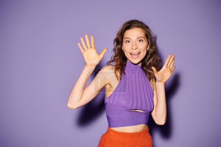 A stylish young woman wears a purple top and an orange skirt against a vibrant purple background.