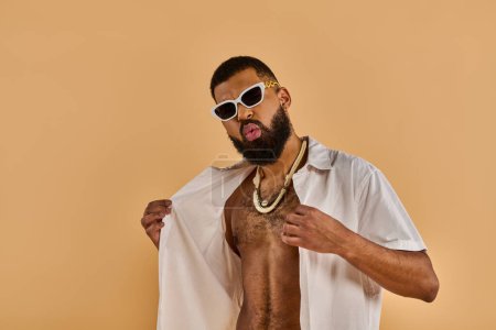 Un homme torse nu avec une barbe élégante porte avec confiance des lunettes de soleil, exsudant une ambiance fraîche et robuste alors qu'il se tient au soleil.