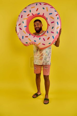 A man playfully holds a giant donut in front of his face, covering it completely. The colorful sprinkles contrast with his expression of joy.