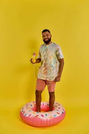 Téléchargez les photos : Un homme en tenue décontractée se tient debout sur un flotteur coloré en forme de donut dans une piscine, tenant un verre dans sa main et profitant du moment. - en image libre de droit