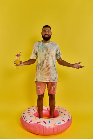 A man confidently balances on top of a giant donut, drink in hand, in a surreal and whimsical scene.