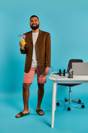 A sophisticated man holds a drink while standing in front of a desk, exuding an air of refinement and relaxation.