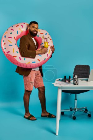 Foto de Un hombre con una sonrisa juguetona sostiene un donut inflable grande delante de un escritorio desordenado, creando una escena caprichosa y surrealista. - Imagen libre de derechos
