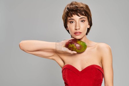 Photo for A captivating young woman stands elegantly in a red dress, holding a vibrant mango. - Royalty Free Image