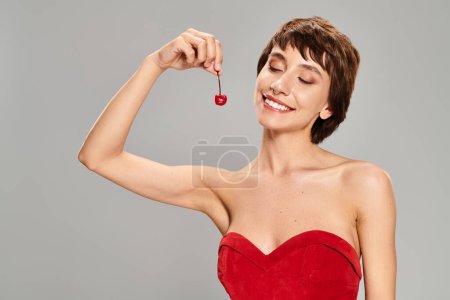 Foto de Mujer joven en un vestido rojo elegante sosteniendo una cereza vibrante. - Imagen libre de derechos