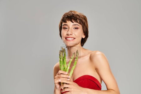A captivating young woman in a radiant red dress holds a bunch of fresh asparagus.