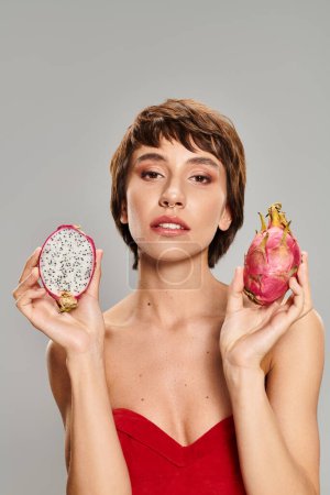 A stunning woman in a red dress gracefully holds a ripe dragon fruit.