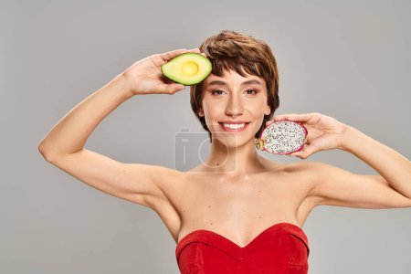 Eine Frau strahlt in einem roten Kleid, während sie eine reife Avocado umklammert.
