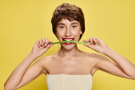 Young woman holds asparagus near mouth, against vibrant backdrop.