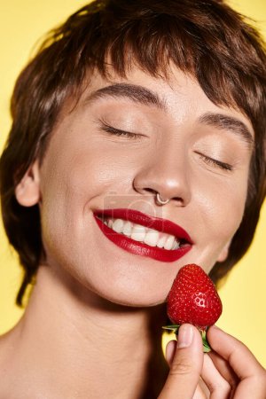 Young woman delicately holds a strawberry to her face against vibrant backdrop.