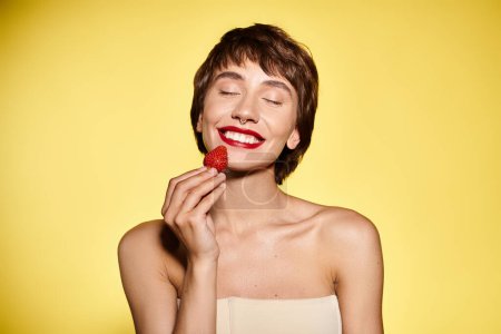 A woman delicately holds a strawberry up to her face against a vivid backdrop.