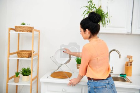 Kobieta w luźnym stroju stoi przy zlewie kuchennym, wykonując prace domowe w pogodny sposób.