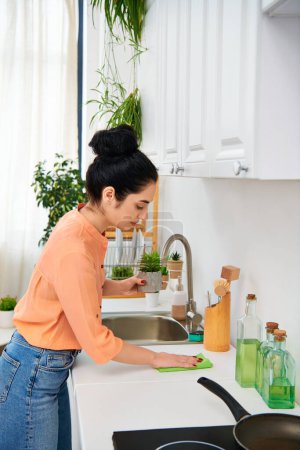Une jeune femme en tenue décontractée nettoie un évier en acier inoxydable dans une cuisine confortable, entourée de savons et de fournitures de nettoyage.