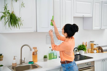A stylish woman in casual attire methodically scrubs the kitchen sink with a vibrant green rag, bringing radiant cleanliness.